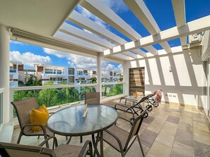 Un patio exterior cubierto con una mesa redonda de vidrio y sillas, un cojín amarillo, y vistas de edificios vecinos.