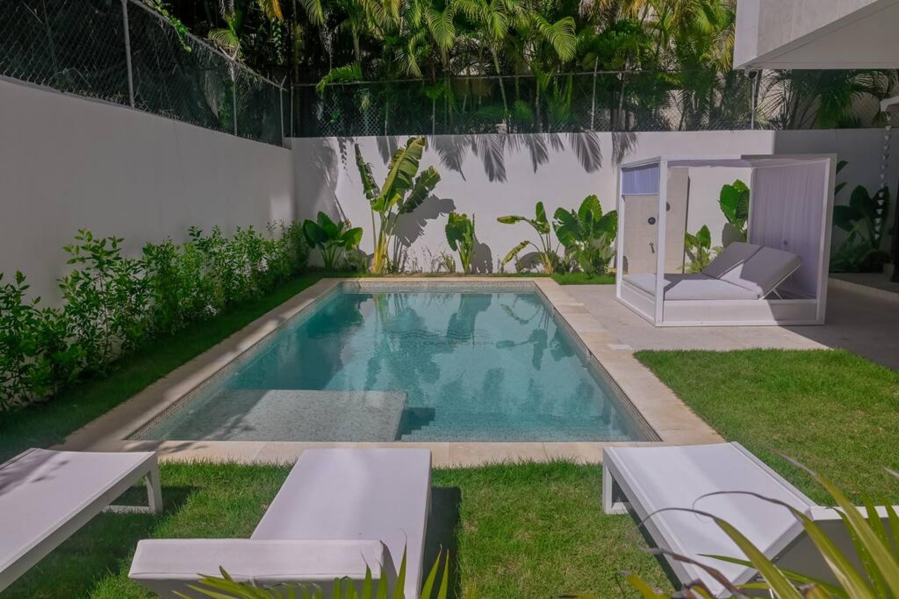 Ein rechteckiger Pool mit weißer Liegefläche, umgeben von üppiger Bepflanzung und einer weißen Wand. Zwei weiße Liegestühle und eine kleine, abgeschlossene Sitzecke mit einer weißen gepolsterten Bank sind vorhanden.