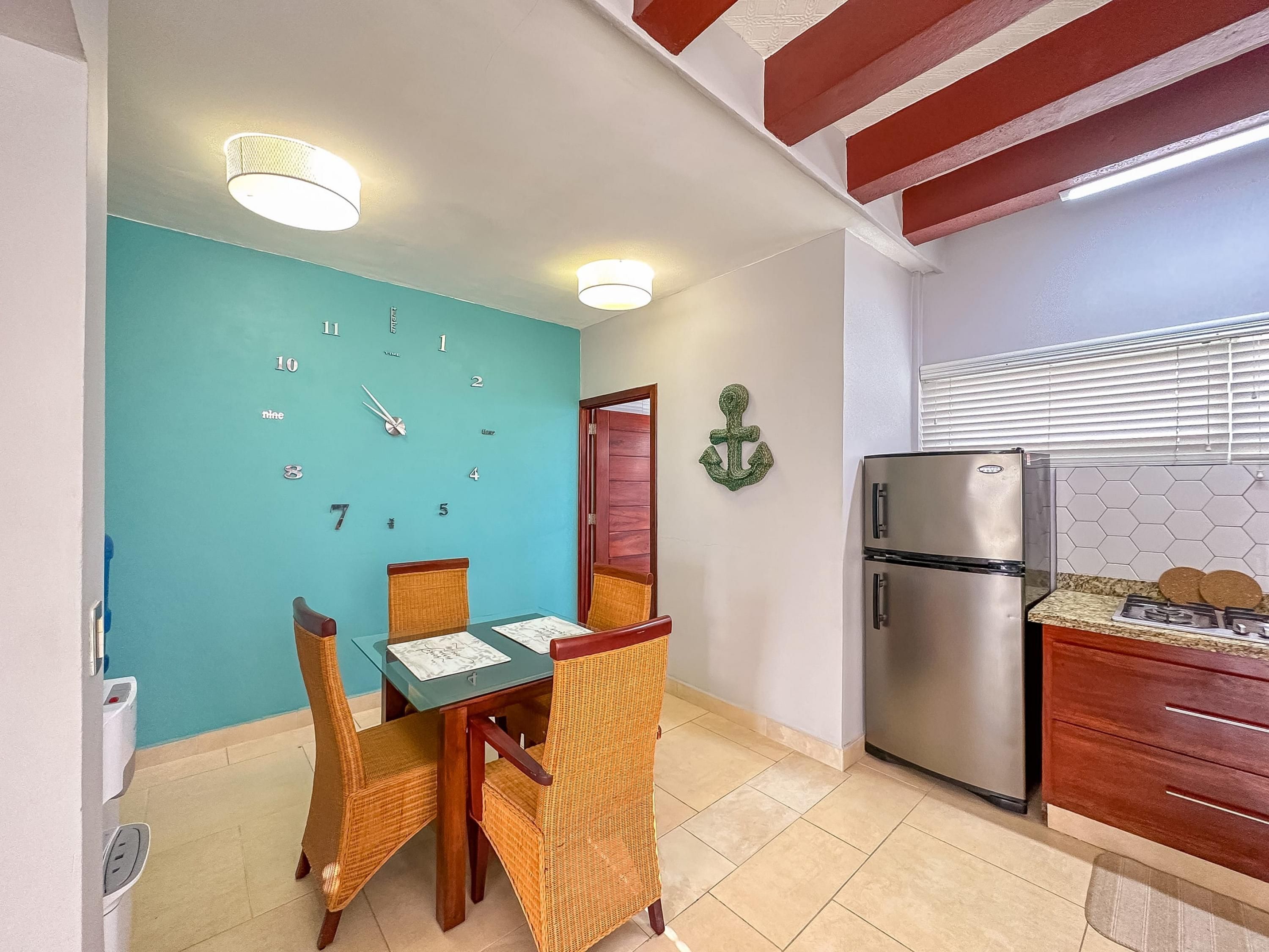 Un comedor con pared color aguamarina, un reloj, sillas de mimbre, una mesa de madera y un refrigerador de acero inoxidable.
