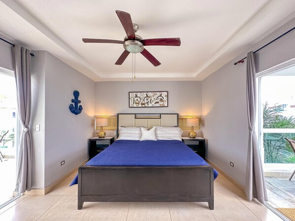 A bedroom with a bed, a ceiling fan, two bedside tables with lamps, a wall-mounted artwork, and a view through a large window. - Incredible 3 BR Penthouse w BBQ Jacuzzi (Punta Cana)