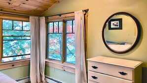 Ein Zimmer mit Holzbalkendecke, einem weißen Schminktisch mit schwarzen Griffen, einem runden Spiegel an der Wand und Fenstern mit Vorhängen.