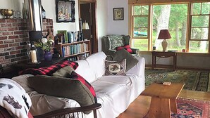 Ein gemütliches Wohnzimmer mit Kamin, Bücherregal, bequemer Couch und Blick durch große Fenster.