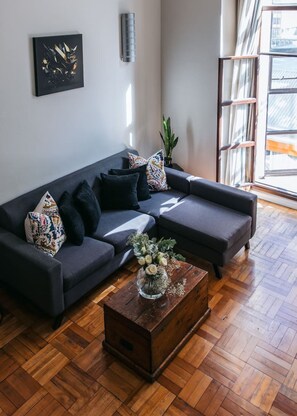 Un salon moderne avec un canapé d’angle gris foncé, une table basse en bois ornée d’un vase de fleurs et une grande fenêtre laissant entrer la lumière naturelle.