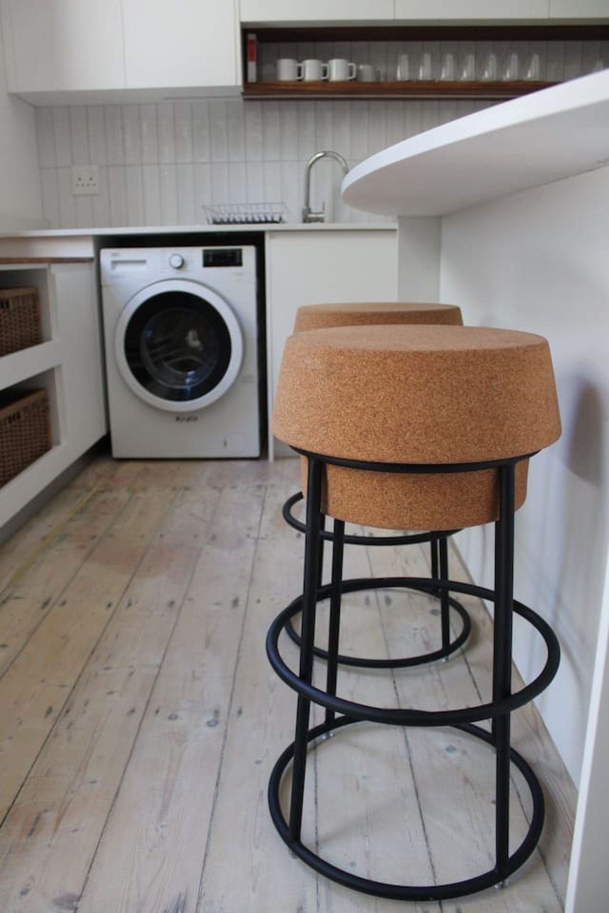 Une cuisine équipée d’un lave-linge, de deux tabourets et d’un plan de travail blanc.