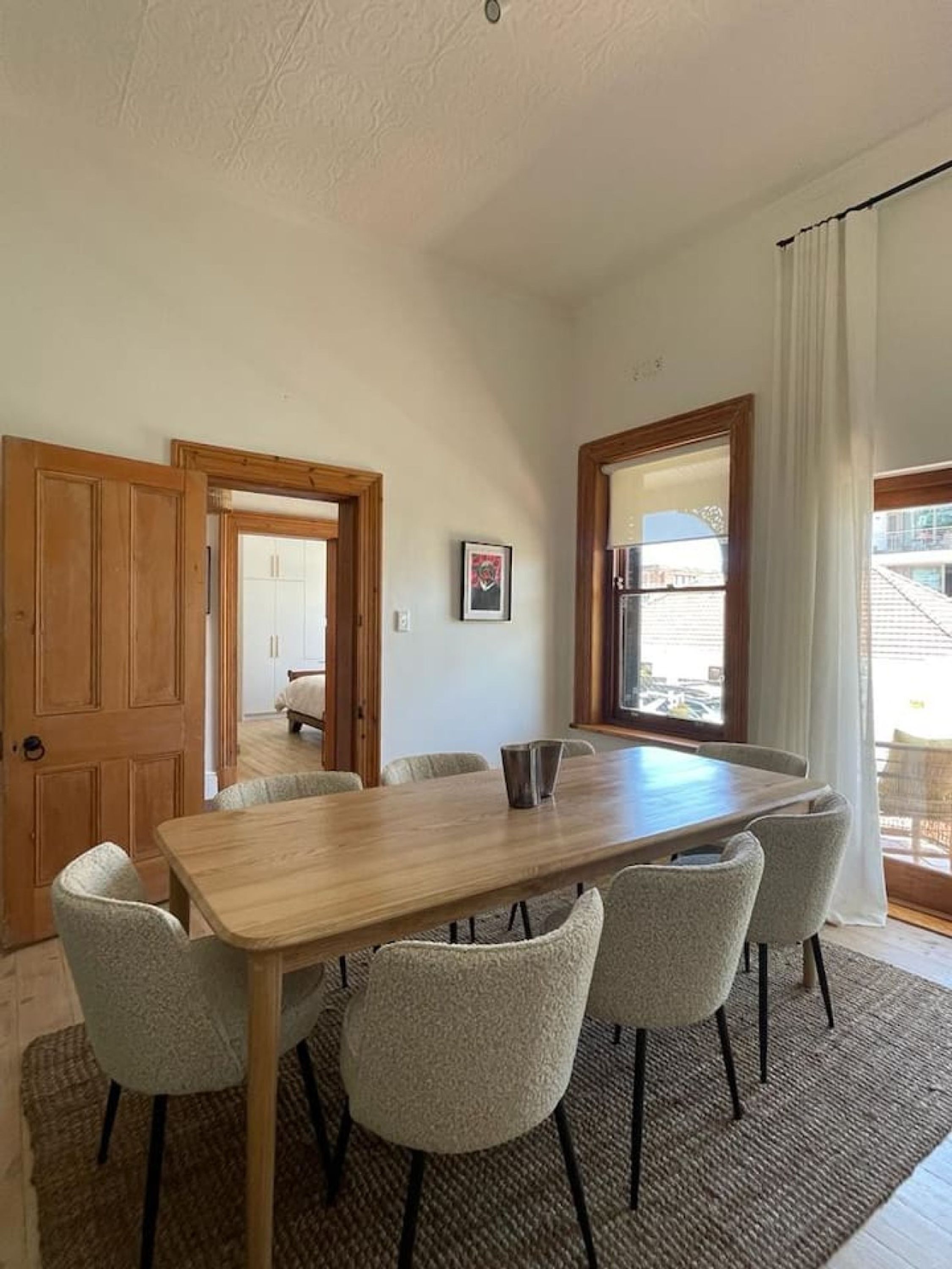 Een eetkamer met een houten tafel en zes stoelen, een lijst met foto aan de muur, en uitzicht door een raam.