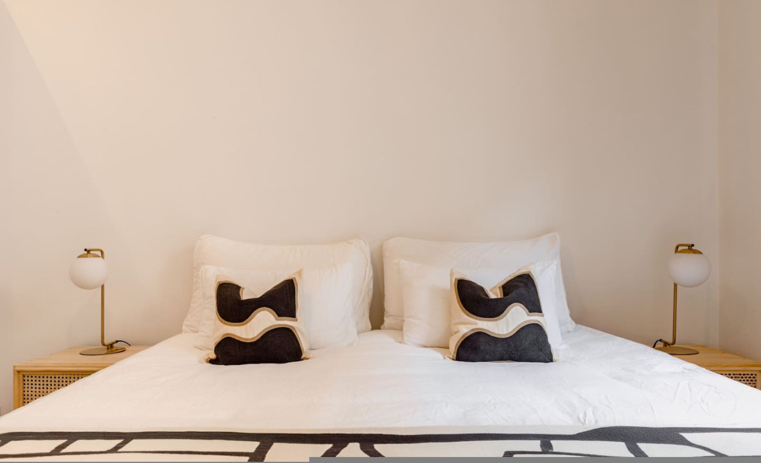Una cama con ropa de cama blanca y dos almohadones decorativos, con dos mesitas de noche y lámparas a cada lado.