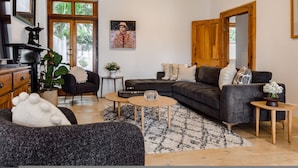 A modern living room with a sectional sofa, a coffee table, and a side table. There is a potted plant and a painting on the wall. - FIR Villa 3 Bedroom House Greenpoint (Cape Town)