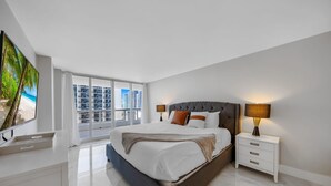 Une chambre moderne avec un grand lit, une table de chevet, un téléviseur et une vue sur la ville grâce à de grandes fenêtres.