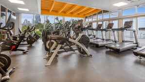 Ein Fitnessstudio mit verschiedenen Cardiogeräten, großen Fenstern und Blick auf Palmen und Gebäude.