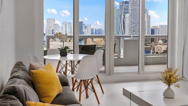Ein modernes Wohnzimmer mit einem grauen Sofa, einem weißen Esstisch mit Stühlen und einem großen Fenster mit Blick auf die Stadt.