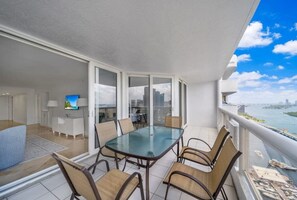 A balcony with a glass table and chairs, a TV, and a view of a city and water. - The Grand Oasis in Miami (Miami)