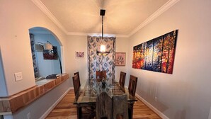 Een eetgedeelte met een tafel met glazen blad, houten stoelen, een gordijn met dessin en een groot abstract schilderij aan de muur.