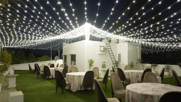 An outdoor dining area with string lights overhead, tables with chairs, and a white building in the background. - Purple Tree Villa Delhi  (New Delhi)