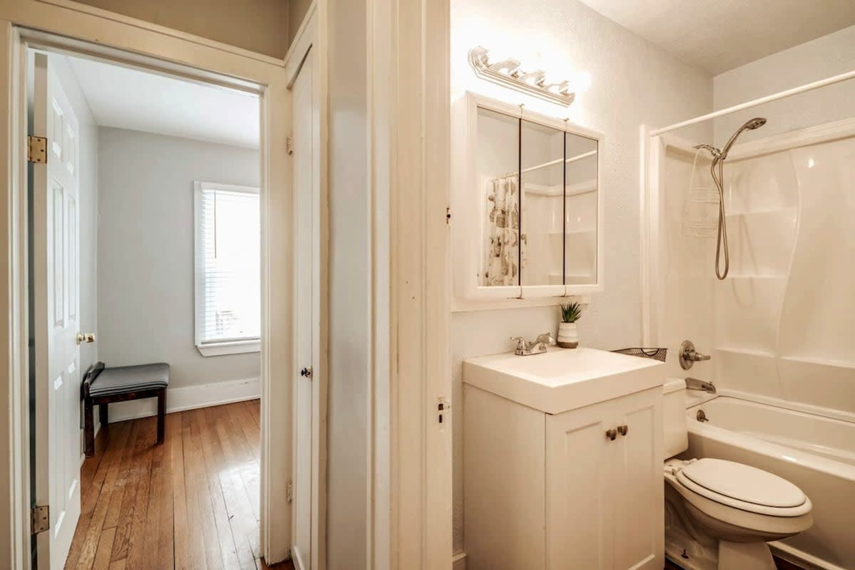 A bathroom with a white sink, mirror, and bathtub. There is a bench in the adjacent hallway.