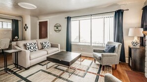 Een woonkamer met een bank, fauteuil, salontafel en een groot raam met gordijnen.