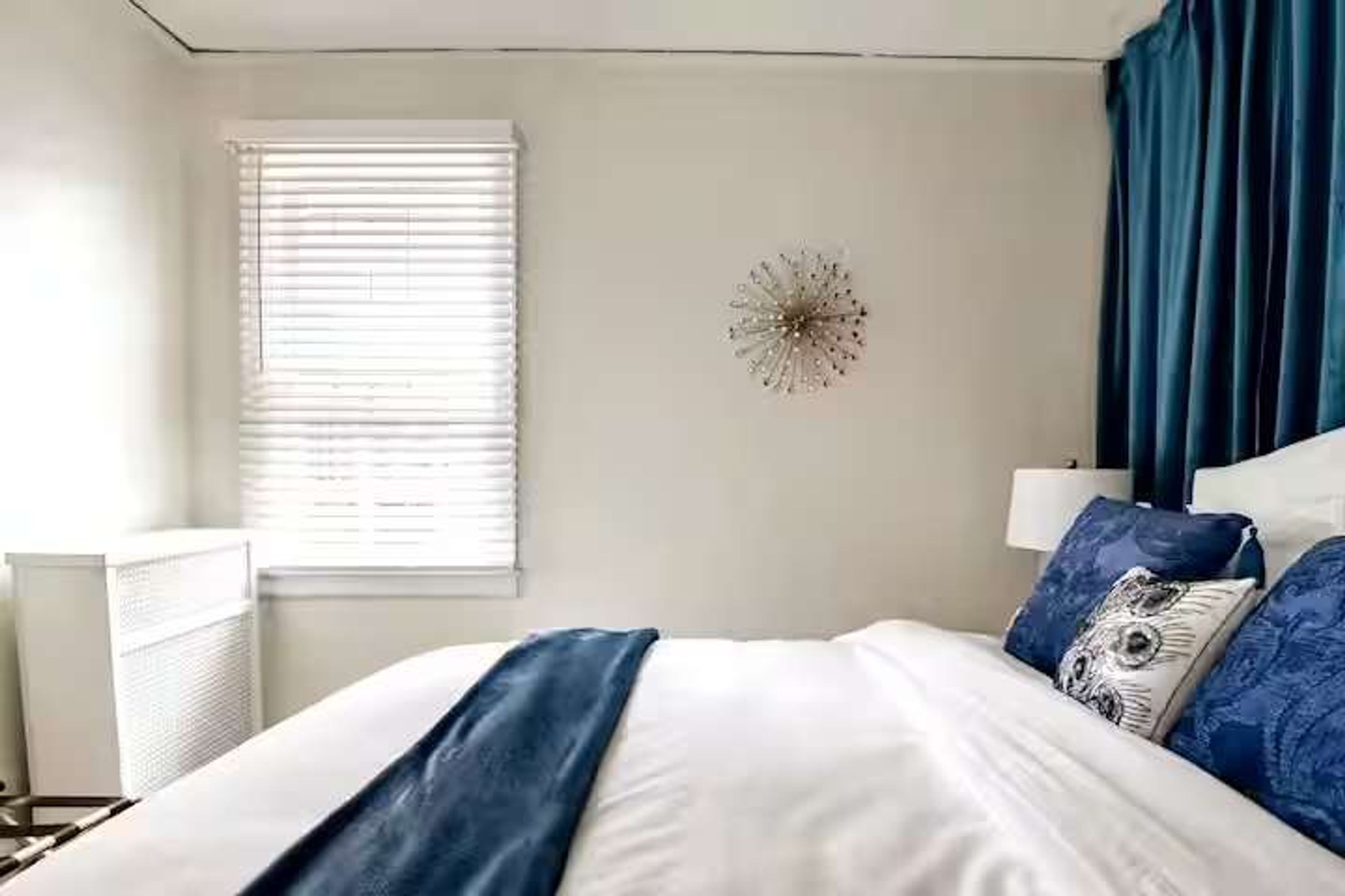 A bedroom with a bed, a window with blinds, a wall clock, and a blue curtain.