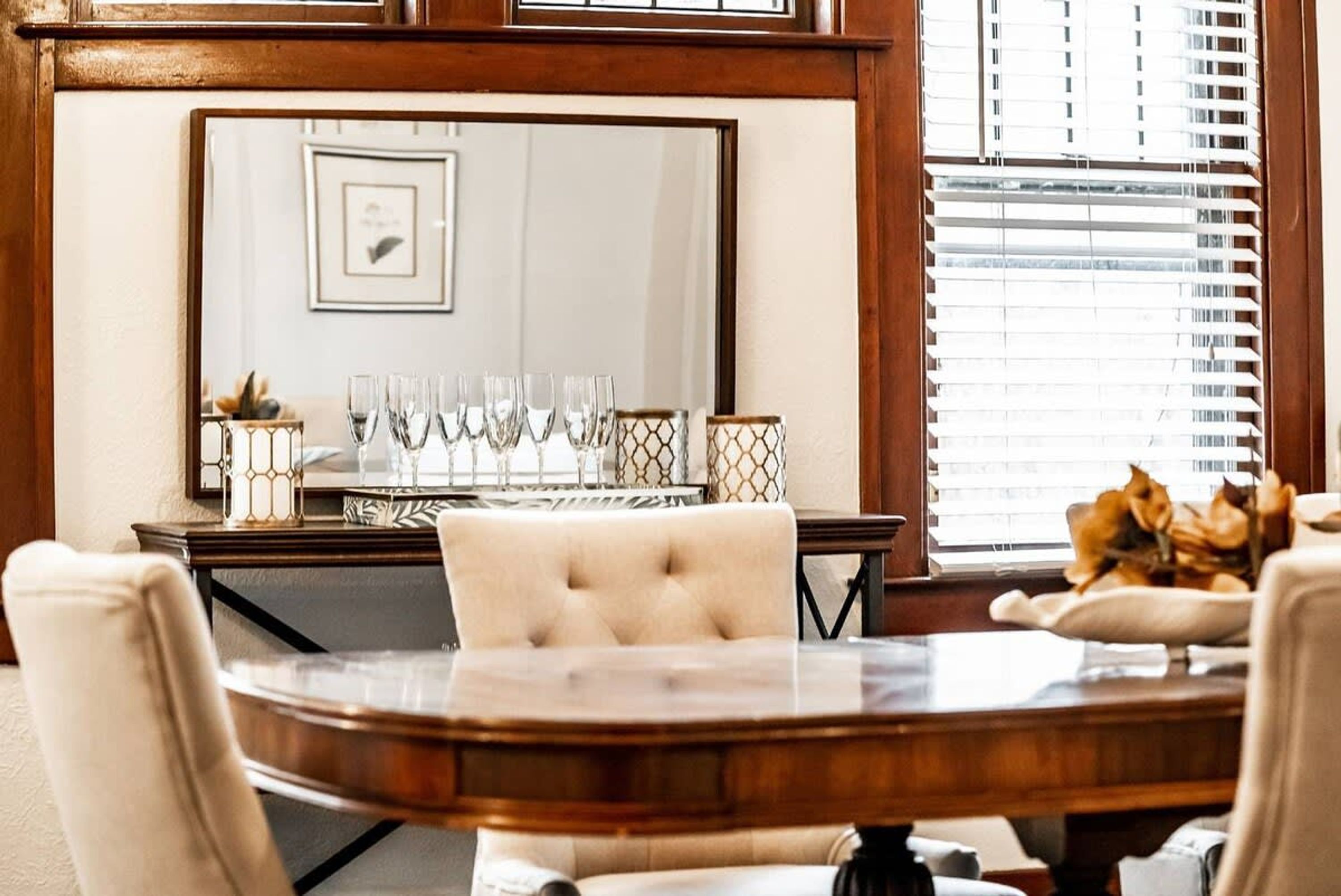 A dining area with a wooden table, white upholstered chairs, and a large mirror above the table.