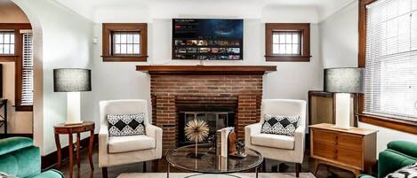 A living room with a brick fireplace, two armchairs, a coffee table, and a TV mounted above the fireplace.