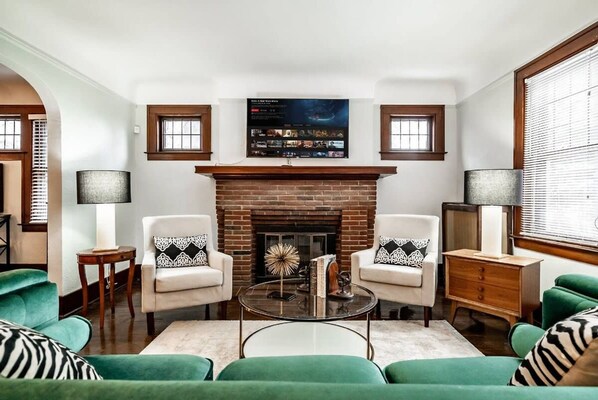 A living room with a brick fireplace, two armchairs, a coffee table, and a TV mounted above the fireplace. - Glamorous Green Beauty in the Heart of University District 15min to Downtown (Detroit)