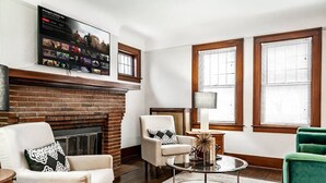 Una sala de estar con chimenea de ladrillo, dos sillones, una mesa de centro redonda y un televisor montado en la pared mostrando una película.