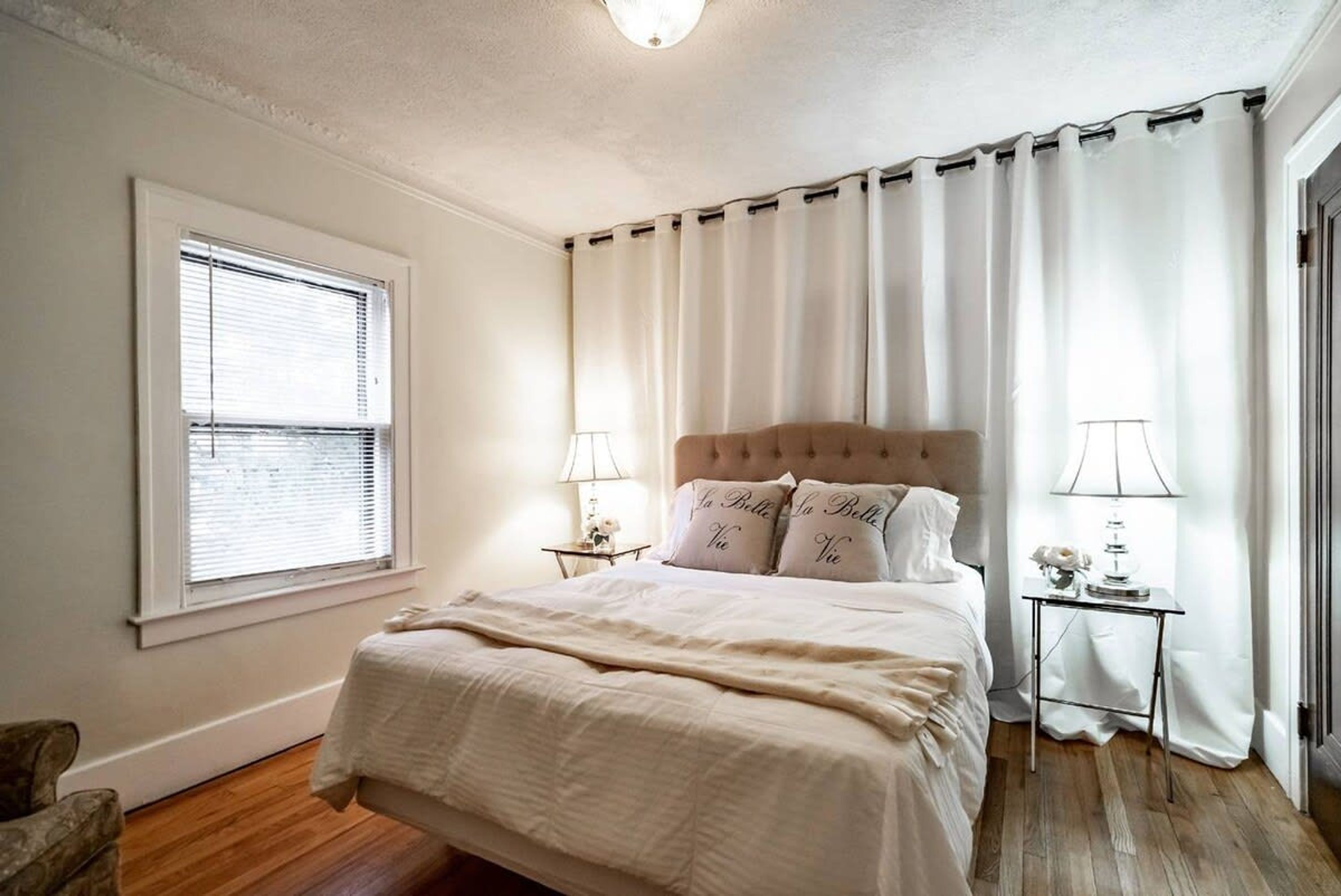 A bedroom with a large bed, two bedside tables with lamps, a window with blinds, and a chair.
