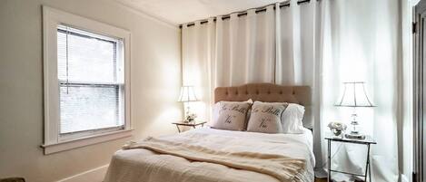 A bedroom with a large bed, two bedside tables with lamps, a window with blinds, and a chair.
