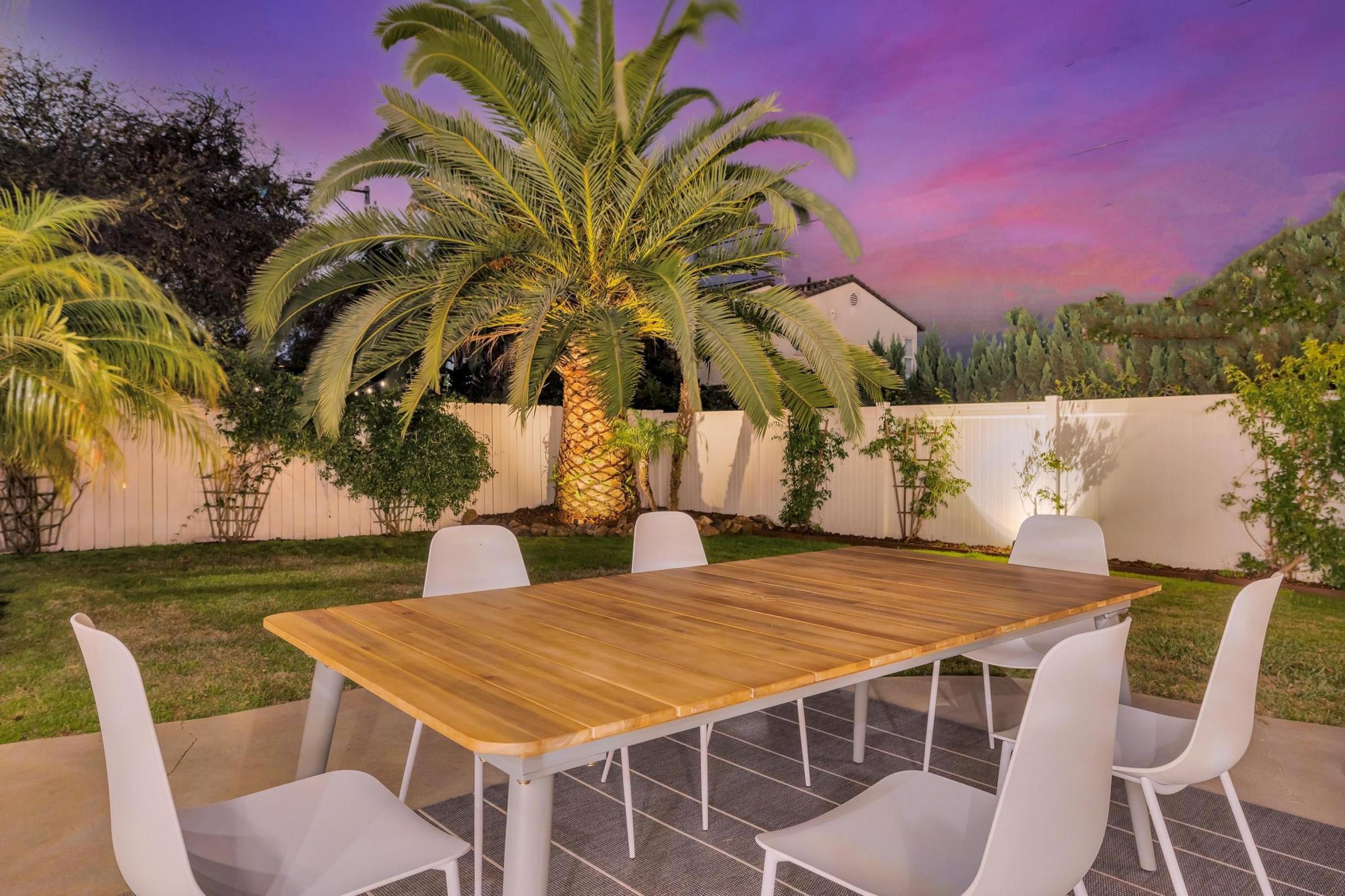 Un patio exterior con una mesa de madera y sillas blancas, rodeado de follaje y una cerca blanca.