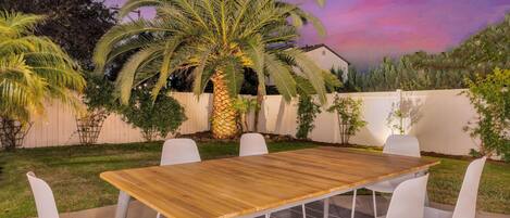 Un patio exterior con una mesa de madera y sillas blancas, rodeado de follaje y una cerca blanca.