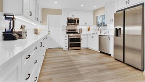 A modern kitchen with white cabinetry, stainless steel appliances, and a wooden floor. - Designer Coastal Paradise w Pool - Steps to Beach (Carlsbad)