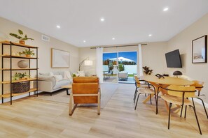 A modern living room with a sofa, a coffee table, and a dining area with chairs and a table. There is a bookshelf with plants and a flat-screen TV mounted on the wall. - Designer Coastal Paradise w Pool - Steps to Beach (Carlsbad)