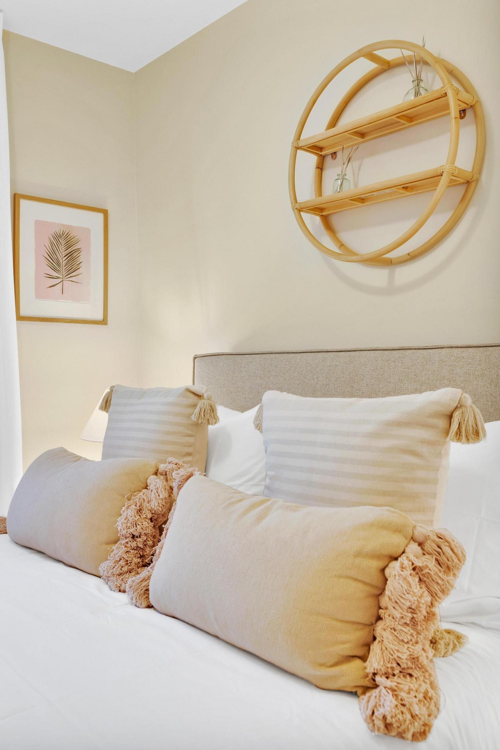 A neatly made bed with white and beige bedding, a round wooden shelf with decorative items, and a framed leaf artwork on the wall.