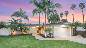 A two-story house with a garage, surrounded by palm trees and a well-maintained lawn. - Designer Coastal Paradise w Pool - Steps to Beach (Carlsbad)