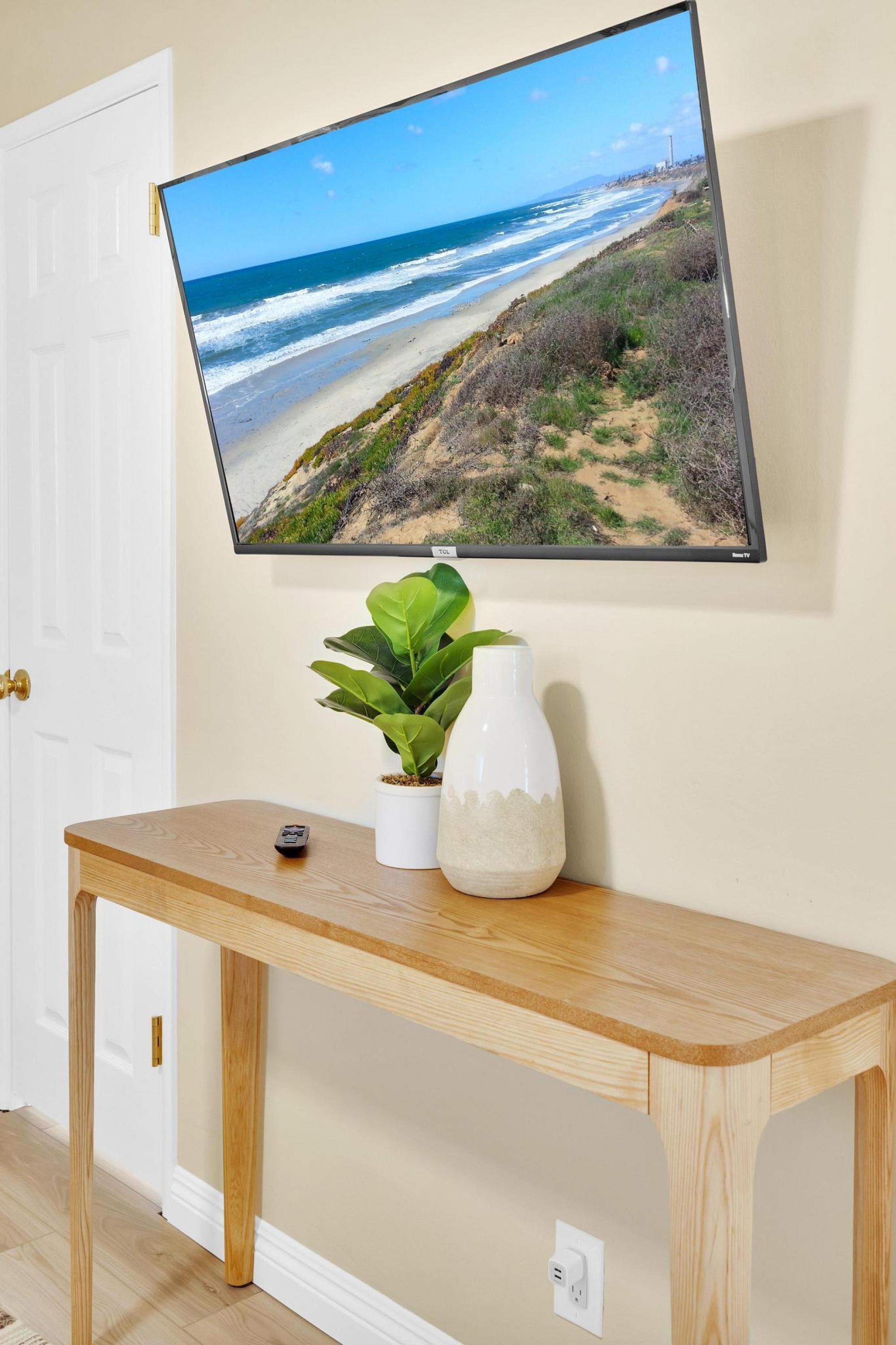 A flat-screen TV mounted on a wall displaying a beach scene, a wooden console table with a potted plant and a vase, and a remote control on the table.
