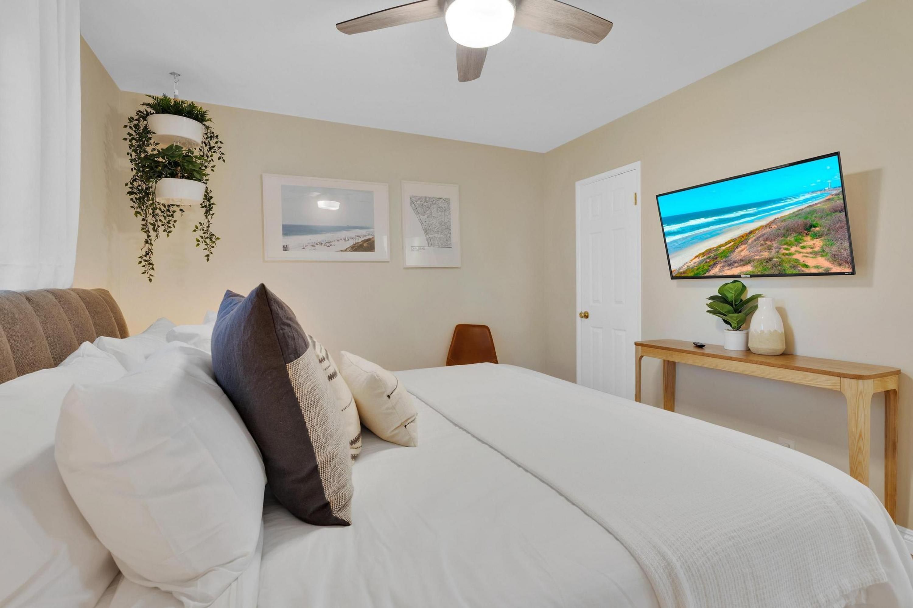 A bedroom with a bed, pillows, a ceiling fan, a wall-mounted TV, and hanging plants.
