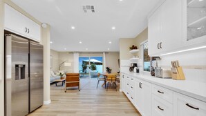 A modern kitchen with white cabinets, stainless steel appliances, and a wooden dining area with a view of the outdoors. - Designer Coastal Paradise w Pool - Steps to Beach (Carlsbad)