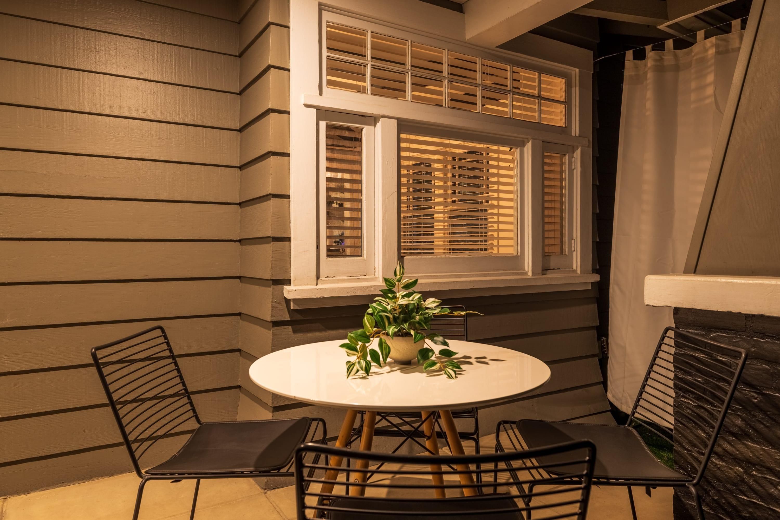 A corner patio with a round table and two chairs, a potted plant, and a window with blinds.