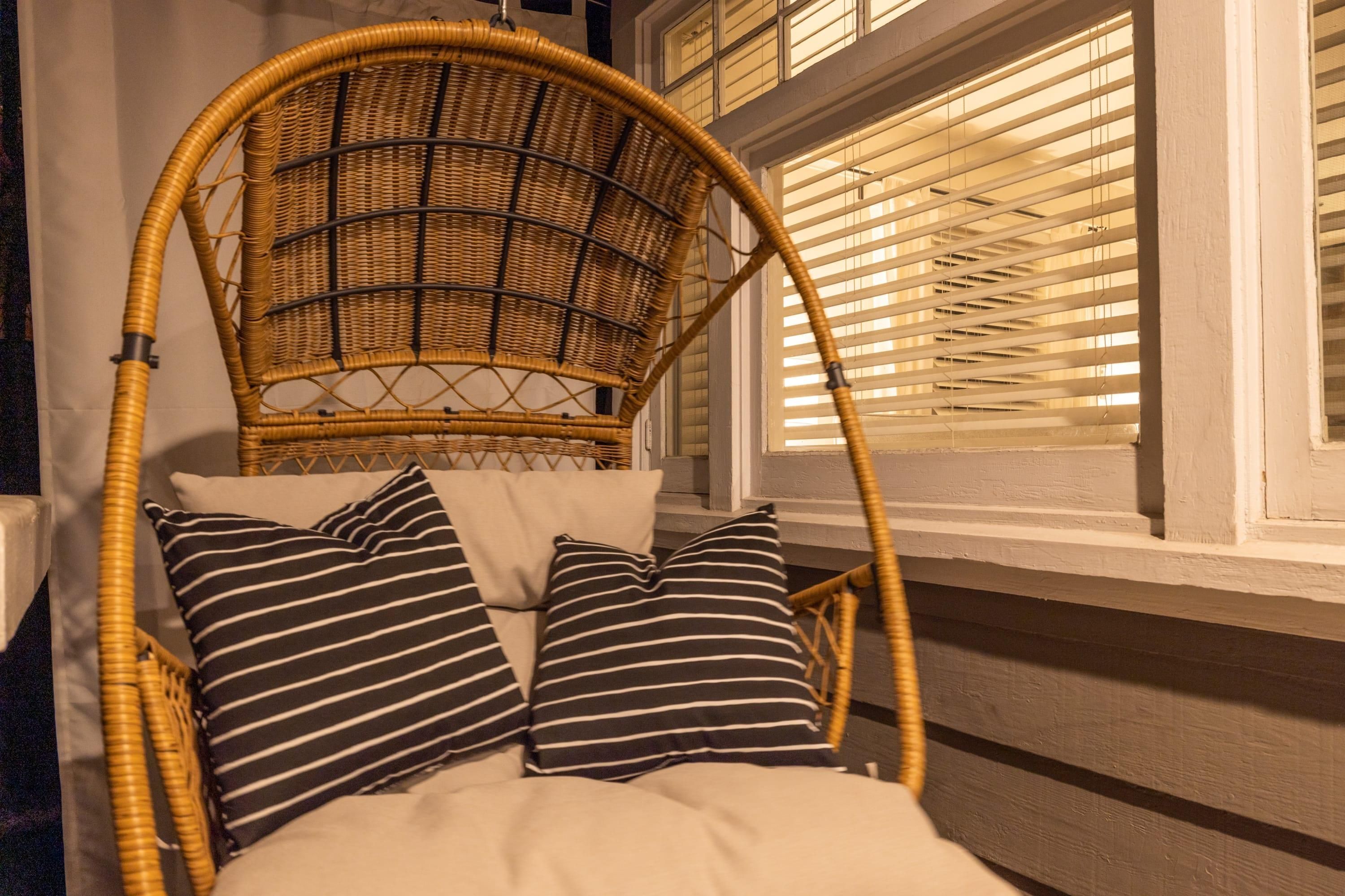 A wicker chair with a cushioned seat and back, positioned in front of a window with blinds.