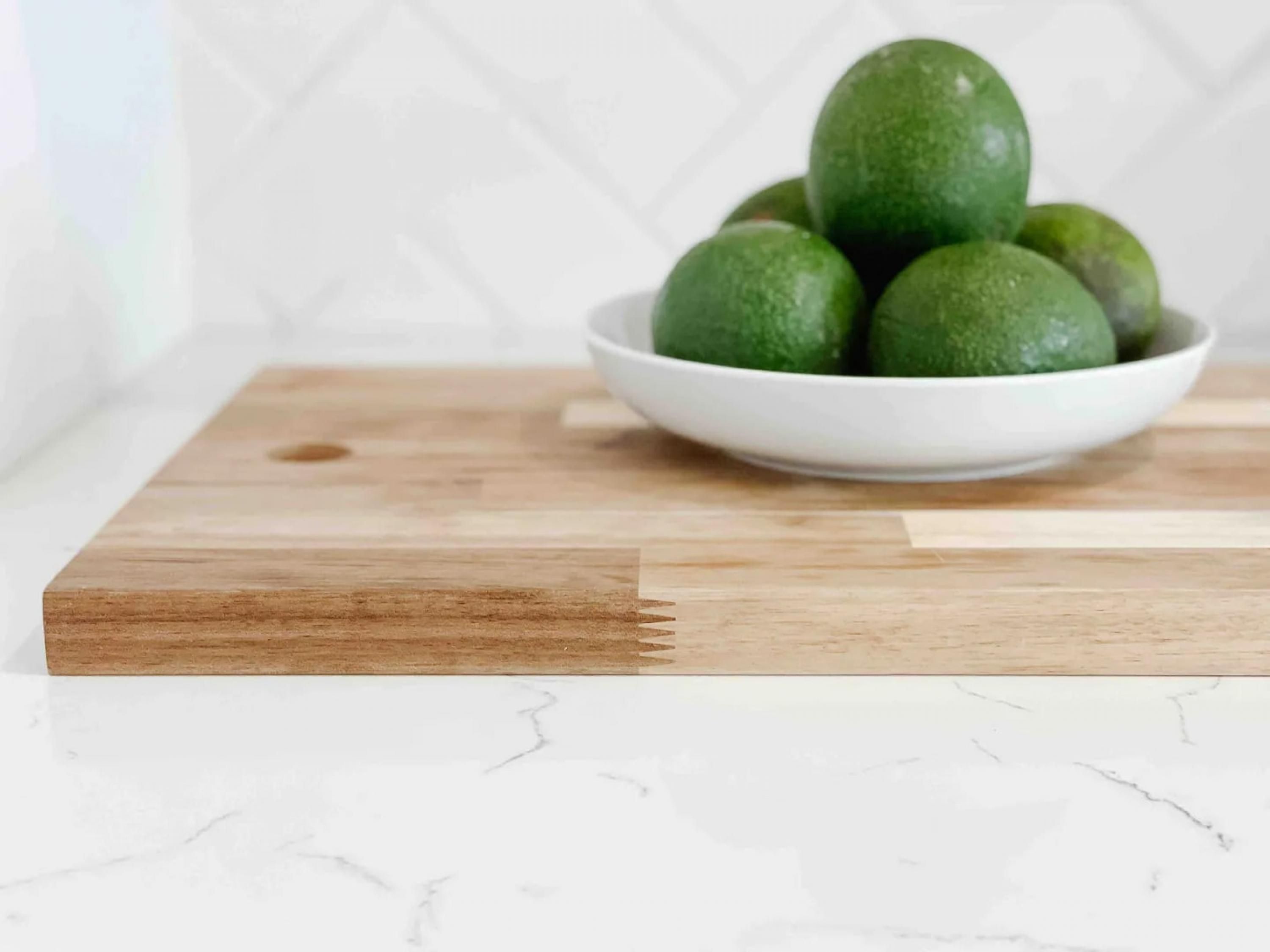 A white bowl with green limes on a wooden cutting board on a marble countertop.