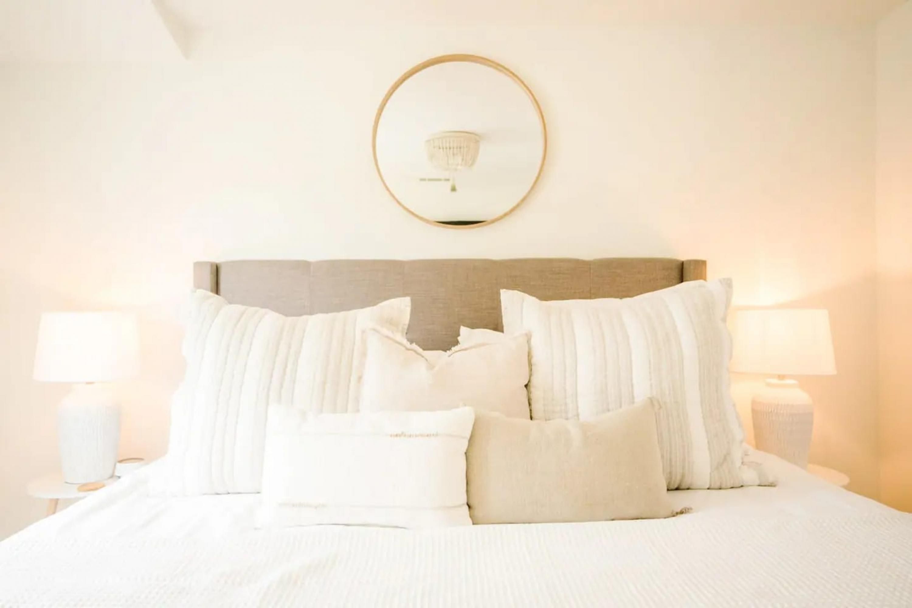 A neatly made bed with white bedding and pillows, a round mirror on the wall, and two bedside lamps.