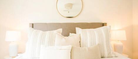 A neatly made bed with white bedding and pillows, a round mirror on the wall, and two bedside lamps.