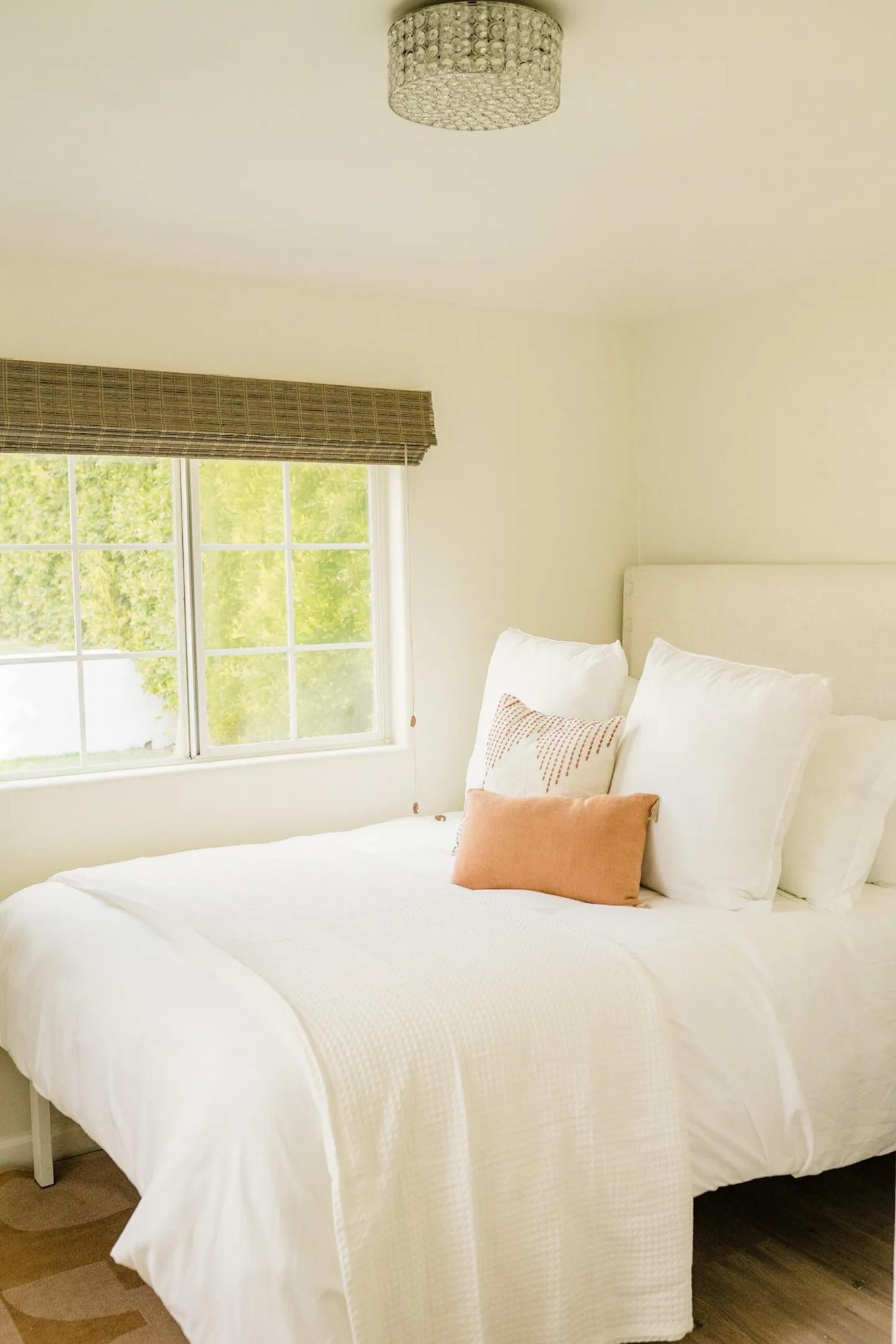 A bedroom with a large bed, white bedding, a window with a view of greenery, and a chandelier.