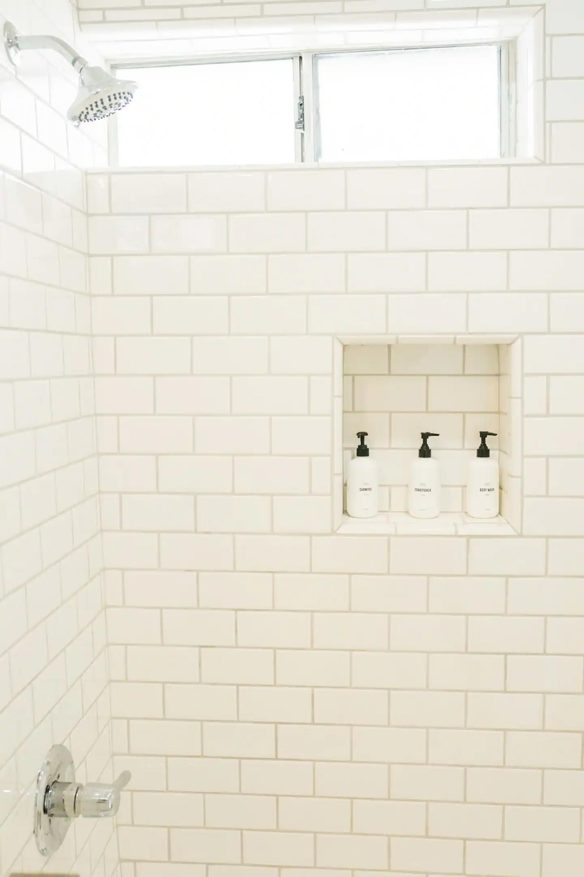 Une salle de bain avec des murs carrelés de blanc, une pomme de douche et une étagère sur laquelle sont posées trois bouteilles.