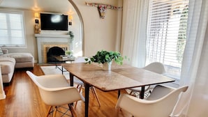 A modern living room with a wooden dining table, white chairs, a flat-screen TV, a fireplace, and a large window with blinds.