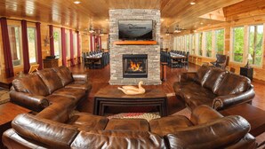 A spacious lounge area with leather sofas arranged around a central fireplace, a mounted television, and a dining area with tables and chairs. - Greenbrier Estate Luxury Theaters Mesmerizing Views (Sevierville)