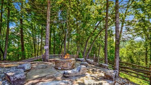 A stone fire pit surrounded by large rocks and a wooden deck in a forested area. - Greenbrier Estate Luxury Theaters Mesmerizing Views (Sevierville)