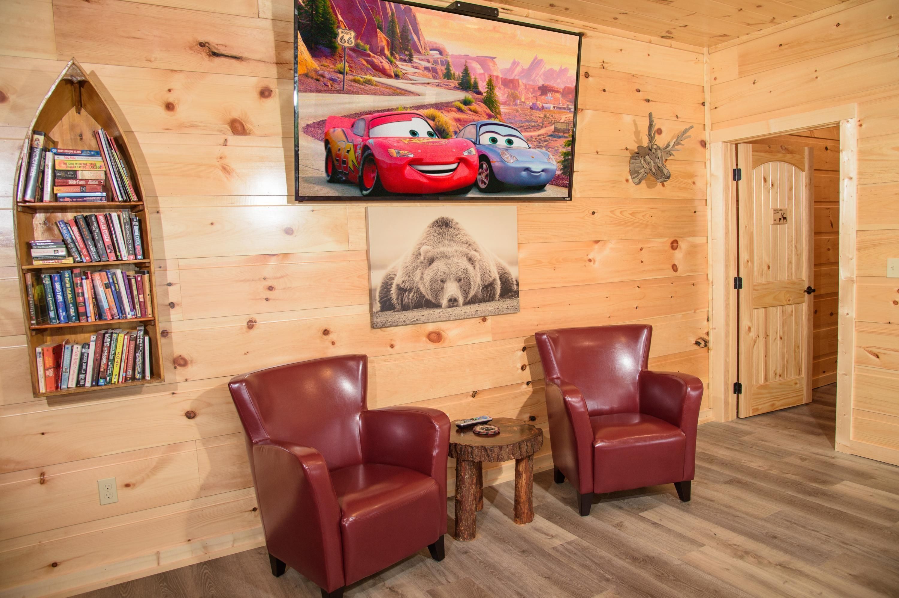A cozy room with wooden walls, a bookshelf, two leather chairs, a small wooden table, and a painting of a bear.