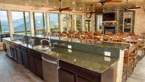 A spacious kitchen with granite countertops, wooden cabinets, and a large island. There are ceiling fans, a stone fireplace, and a view of the mountains through large windows. - Skyline Splendor Luxe Retreat With Breathtaking Views Over Downtown Gatlinburg (Gatlinburg)