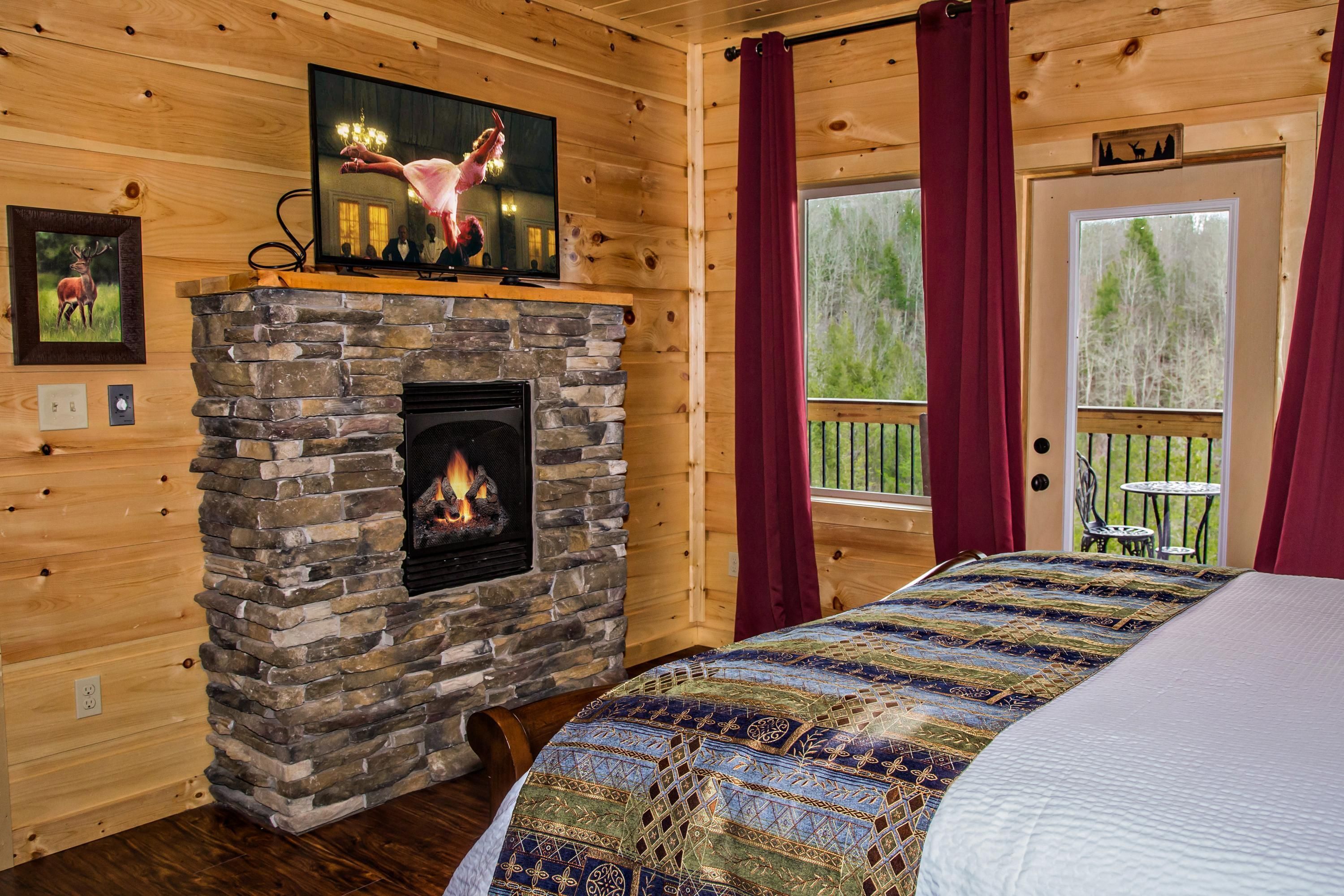 Una habitación de cabaña acogedora con chimenea de piedra, colcha acolchada, paredes de madera y un ventanal con cortinas.