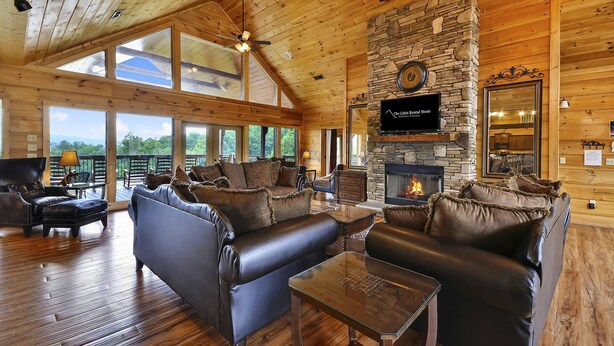 A spacious cabin interior with a stone fireplace, leather sofas, and a large window overlooking a scenic landscape. - Gatlinburg s Finest Majestic Views Home Theater Hot Tubs and More (Gatlinburg)
