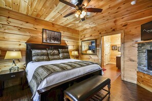 Une chambre à coucher située dans une cabane en bois, comprenant un lit, un banc, une table de chevet, une lampe, un ventilateur de plafond et une cheminée en pierre.